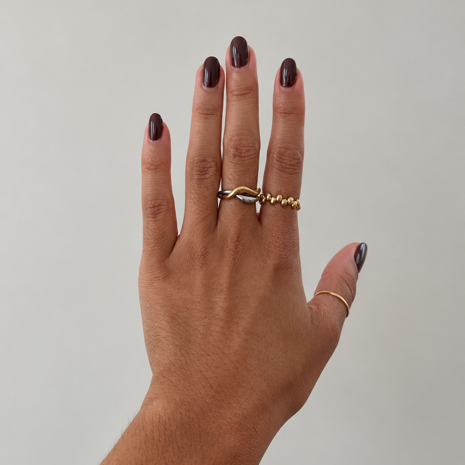 A hand with dark brown manicured nails wearing three gold rings, including a silver-toned wave ring, a beaded gold ring, and a thin gold band, against a plain light gray background.